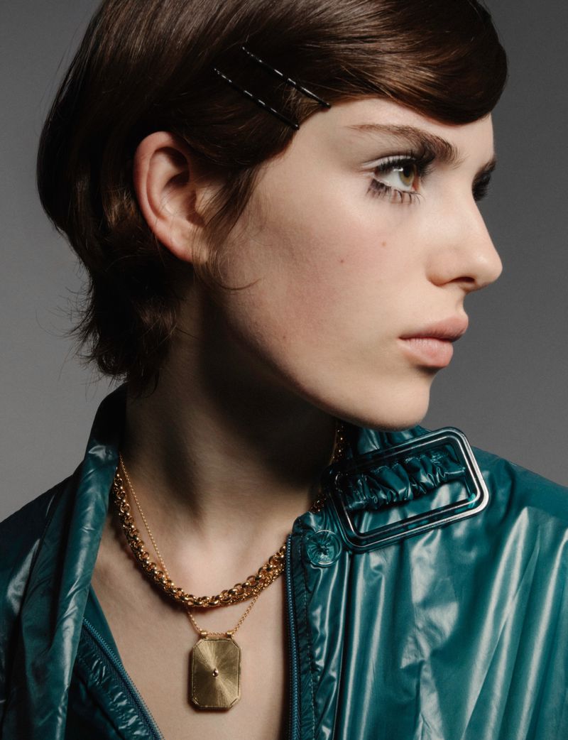 Close-up studio shot of a young adult woman with short brown hair, wearing a turquoise jacket and gold necklaces, and posing in profile.