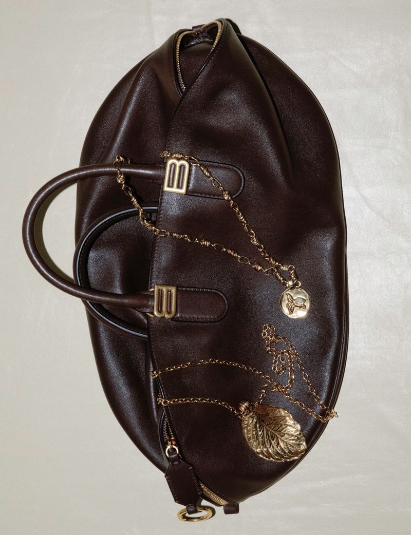 A brown leather satchel-style handbag with a top handle, adorned with two gold-colored pendant necklaces on an off-white surface.