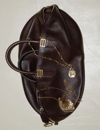 A brown leather satchel-style handbag with a top handle, adorned with two gold-colored pendant necklaces on an off-white surface.