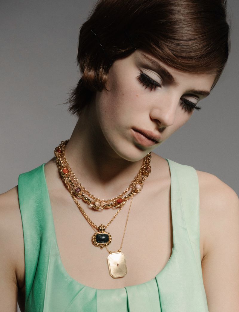Close-up of a fair-skinned woman with dark hair and heavy eye makeup wearing a light green top and layered gold necklaces.