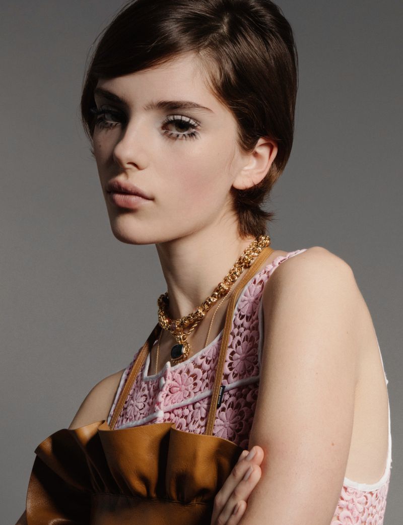 A close-up portrait of a woman with short dark hair wearing a pink lace top, gold chain necklace, and brown ruffled leather accessory.