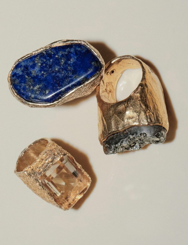 A close-up studio shot features three gold rings adorned with gems: lapis lazuli, a clear gemstone, and an opaque, rough stone.