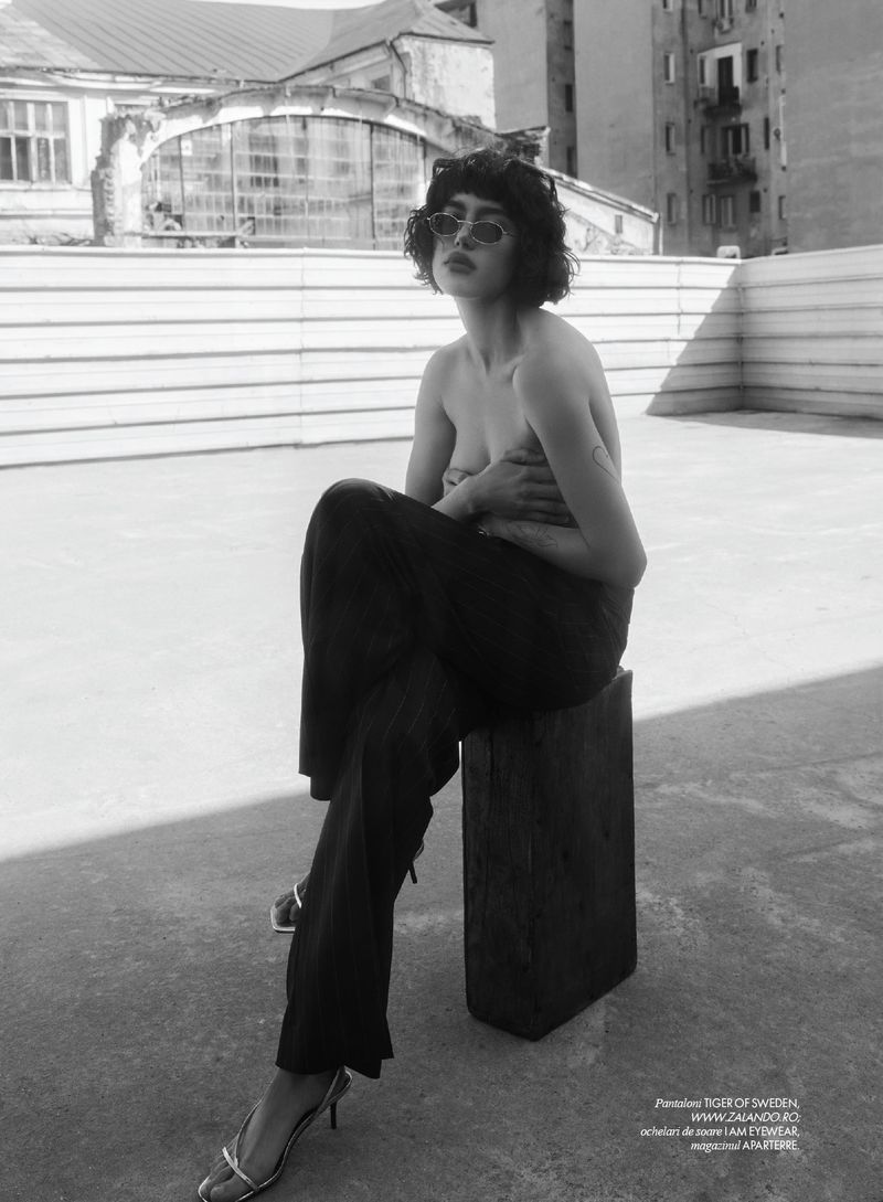 Monochrome portrait of a young adult woman sitting on a wooden block on a rooftop, wearing sunglasses and pants.