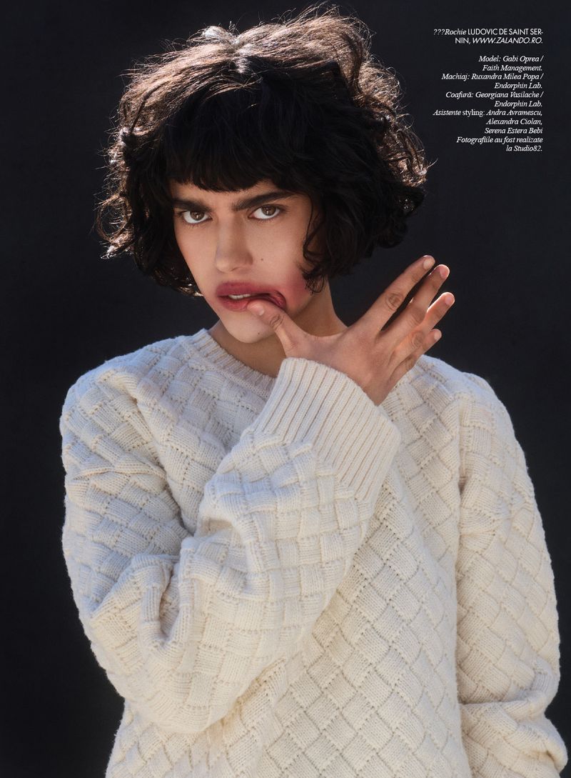 Fashionable female model poses in a studio, wearing a cream-colored textured sweater against a dark backdrop, showcasing contemporary style.