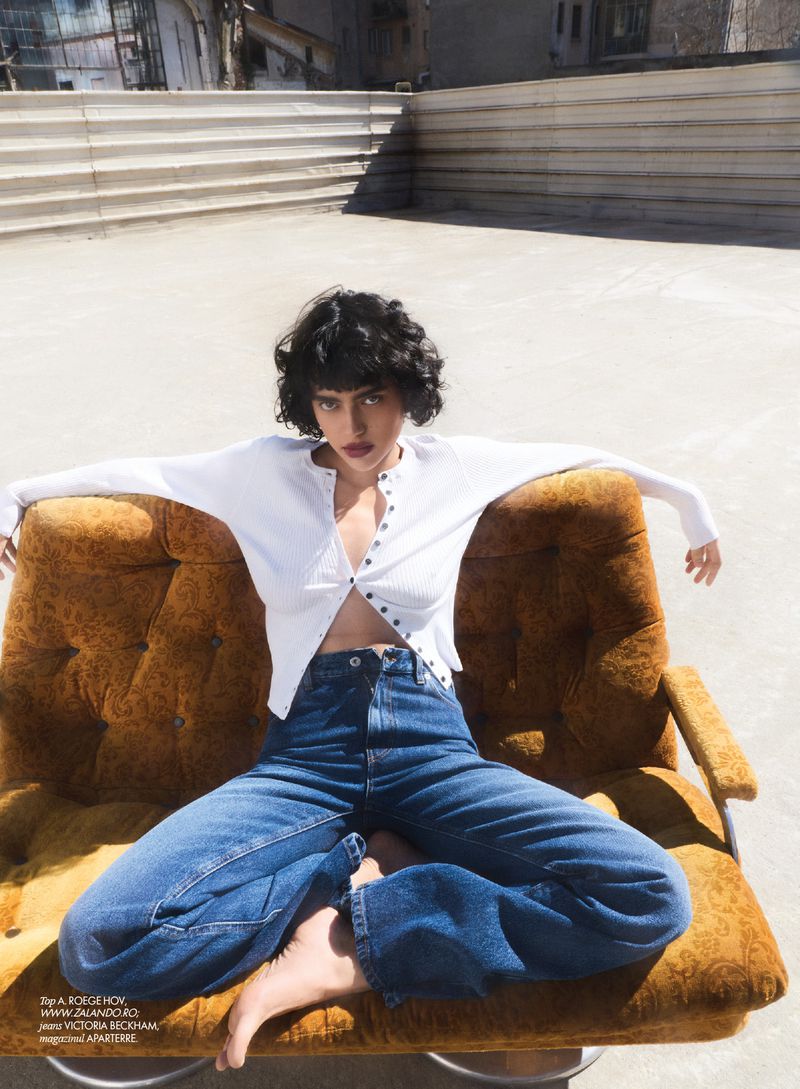 Fashion shot of a woman in a white top and jeans sitting in an orange chair on a rooftop with a blurred urban background, stylish and relaxed.