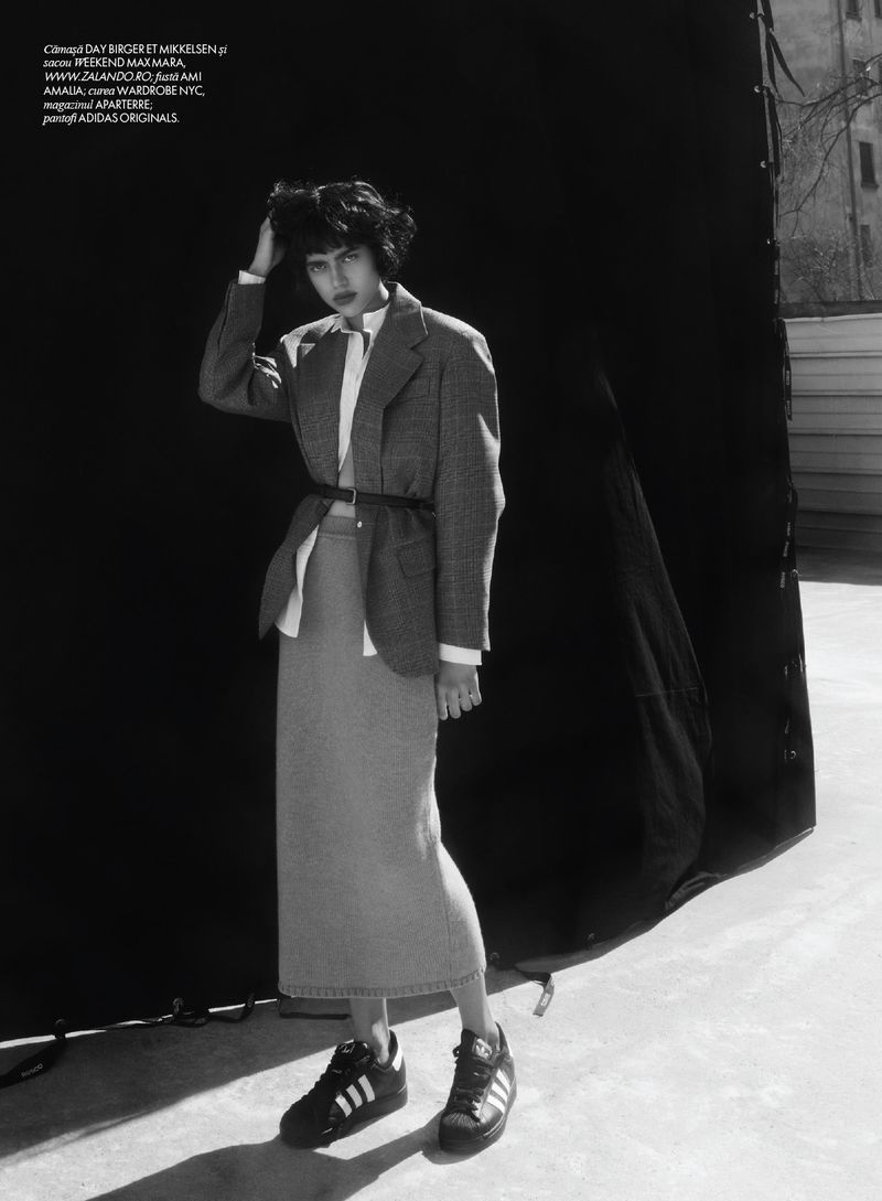 Model with dark short hair wearing an oversized grey blazer and midi skirt with black sneakers against a black backdrop
