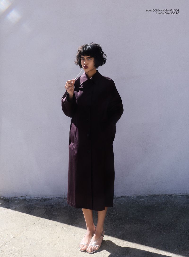 A woman in a dark coat and clear shoes stands in front of a white wall in an outdoor setting with natural light and shadows.