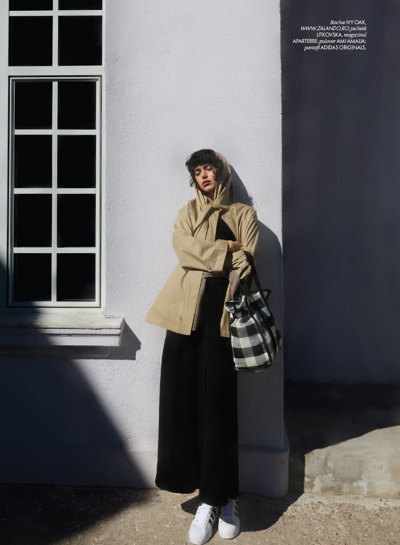 Young woman in beige trench coat, black wide-leg pants, and white sneakers stands outdoors against a building with window.