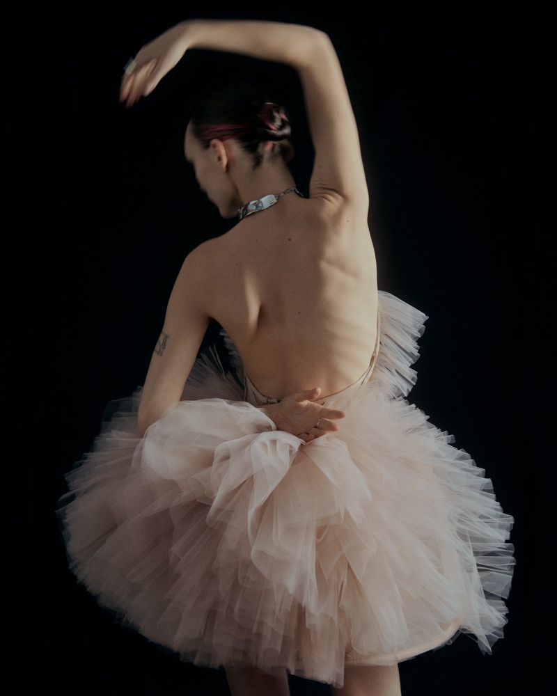 Ballet dancer in pink tutu poses with arms overhead against a black background in a studio setting, captured in a medium shot.