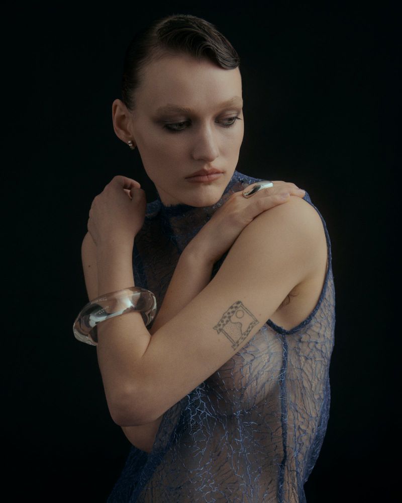 Portrait of a woman wearing a sheer blue textured top, large clear bangle, and silver ring against a dark background