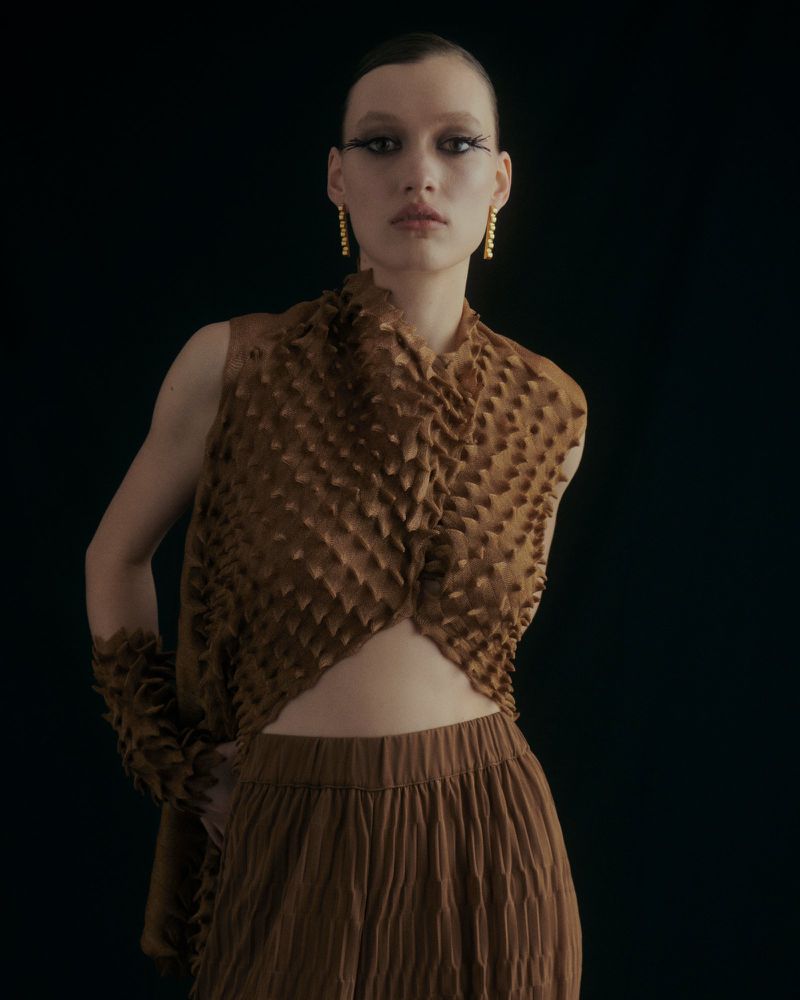 Fashion portrait of a young adult woman with unique eye makeup posing in a brown textured vest and skirt against a dark backdrop.