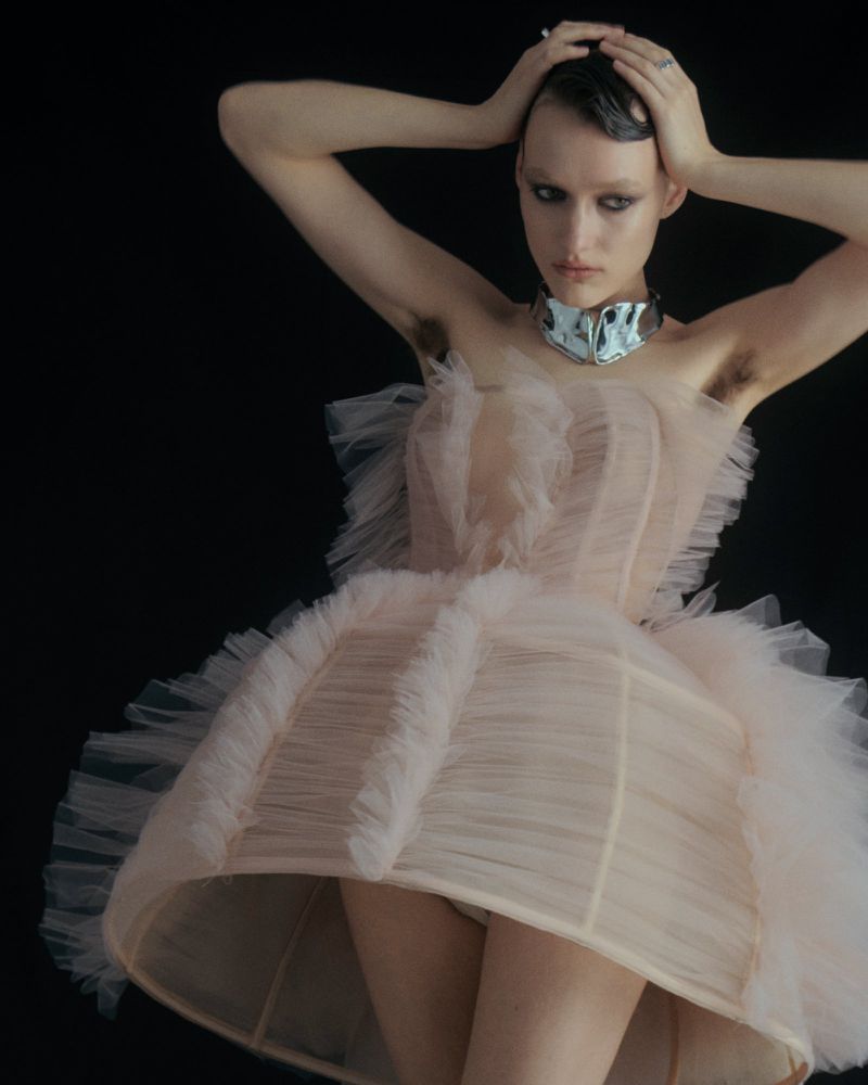 Young adult woman in a pink tulle dress poses in a studio, hands behind her head, silver choker, short hair, on a black background.
