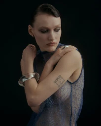 Portrait of a woman wearing a sheer blue textured top, large clear bangle, and silver ring against a dark background