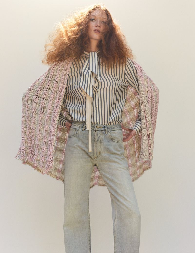 Fashion photo of a red-haired model wearing a striped shirt, jeans, and a crocheted shawl, posed against a neutral studio backdrop.
