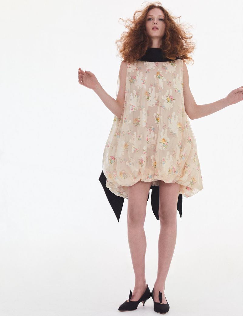 A red-haired fashion model poses in a floral-print mini-dress with black accents against a white backdrop, completed with black kitten heels.