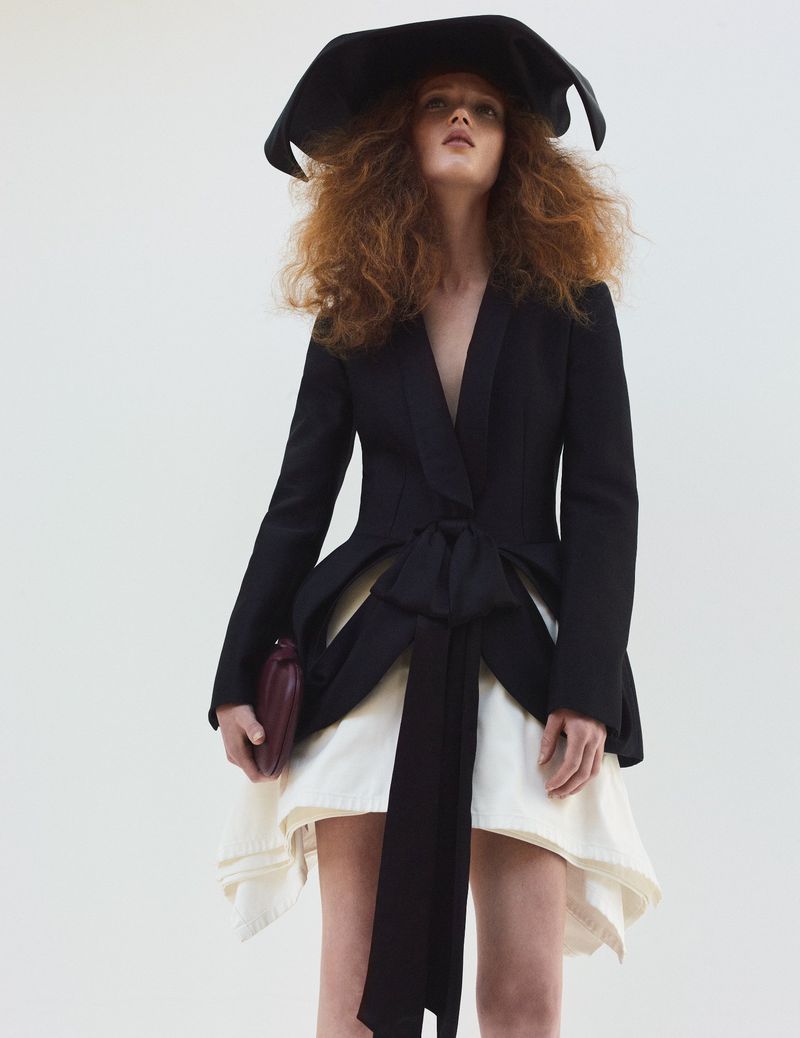 A redhead fashion model wears a black blazer with a white dress and a wide-brimmed hat against a neutral backdrop in a studio setting.