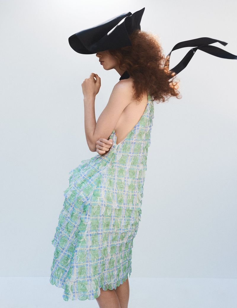 Fashion shot of a person wearing a patterned dress and a large black hat with ribbons against a plain white background in a studio.