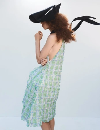 Fashion shot of a person wearing a patterned dress and a large black hat with ribbons against a plain white background in a studio.