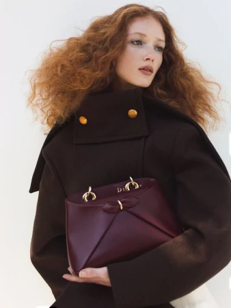 A woman with red hair poses while holding a burgundy leather handbag and wearing a dark brown wool coat with golden buttons.