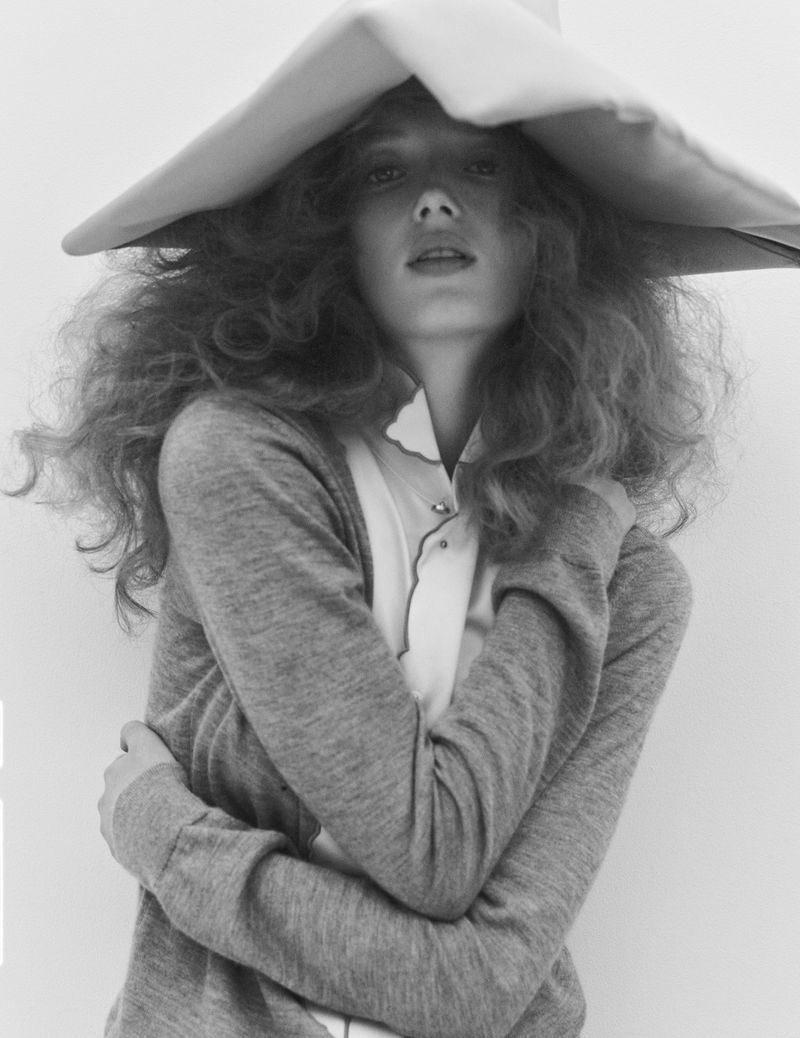 Monochrome portrait of a young woman with curly red hair and a large, wide-brimmed hat, wearing a grey sweater against a neutral background.