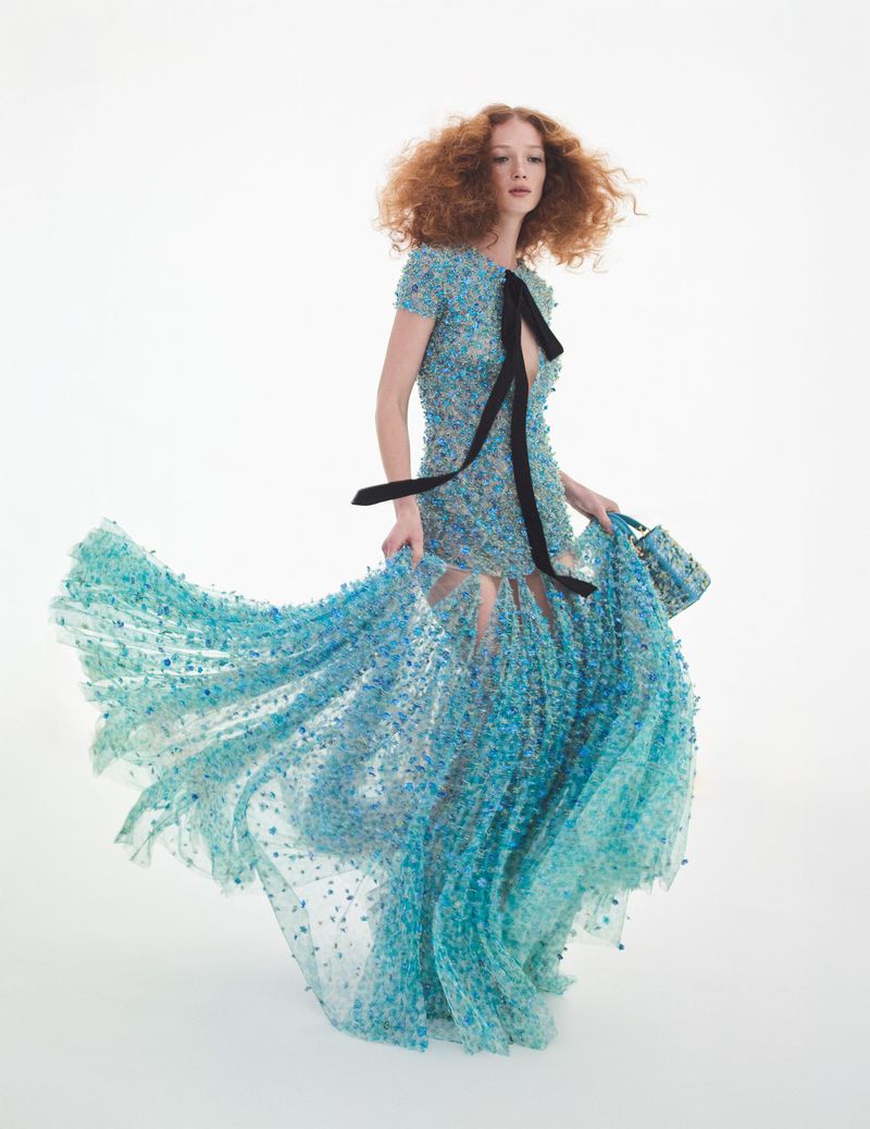 A fashion image features a red-haired woman in a blue beaded gown, posing in a dynamic stance against a bright white backdrop in a studio.