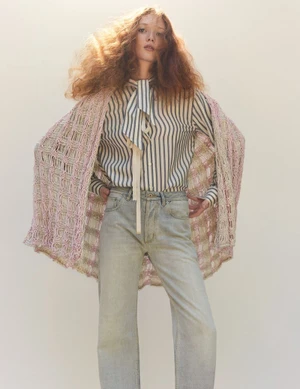 Red-haired female model wearing a striped shirt, jeans, and a knitted poncho against a plain background