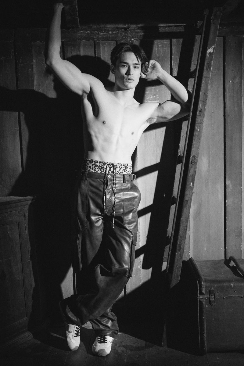 Black and white photo of muscular Asian man in leather pants leaning against ladder in dim indoor light. Fashion editorial photo.