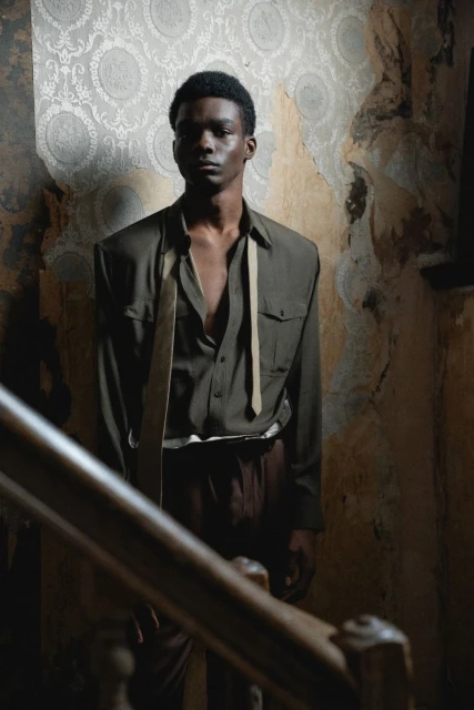 Portrait of a young black male standing on a staircase in a decayed interior. He is wearing a shirt and trousers with suspenders.