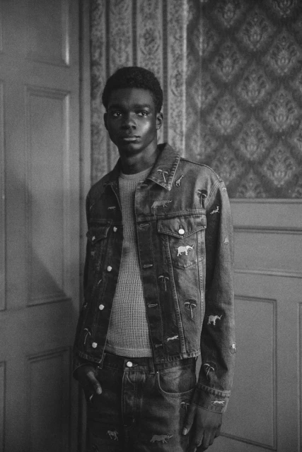 Monochrome portrait of a Black male model in a denim jacket and jeans with animal embroidery, standing against an ornate wall indoors.