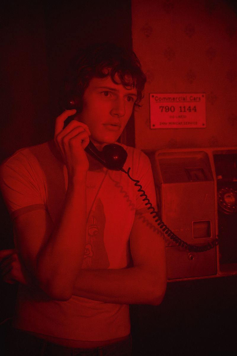 A young adult man with curly dark hair is using a vintage telephone in a red-lit indoor setting with dated payphones in the background.
