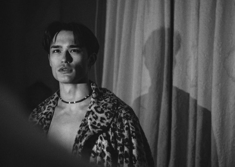 Monochrome portrait of a male model wearing an animal print jacket and beaded choker necklace against a curtained backdrop.