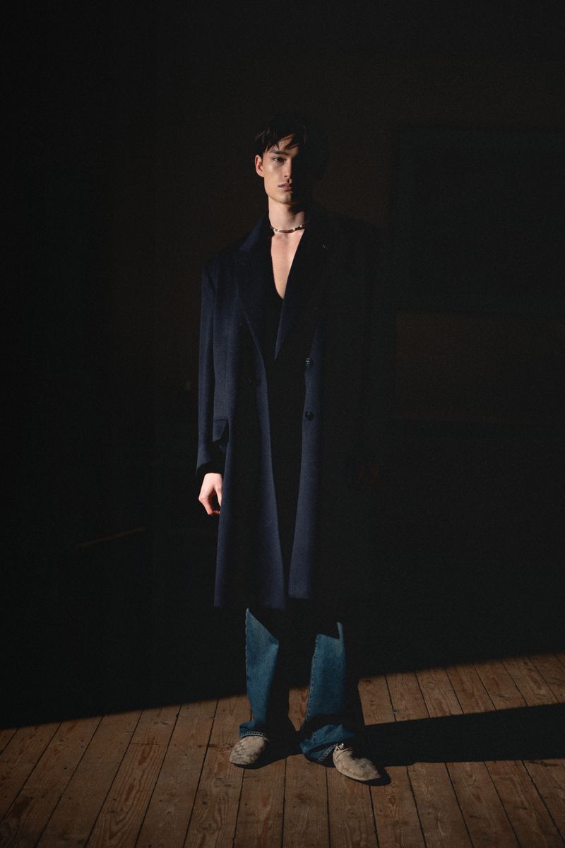 Portrait of a young man in a dark blue overcoat and jeans standing indoors on wooden floorboards in a spotlight.