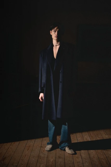 Portrait of a young man in a dark blue overcoat and jeans standing indoors on wooden floorboards in a spotlight.