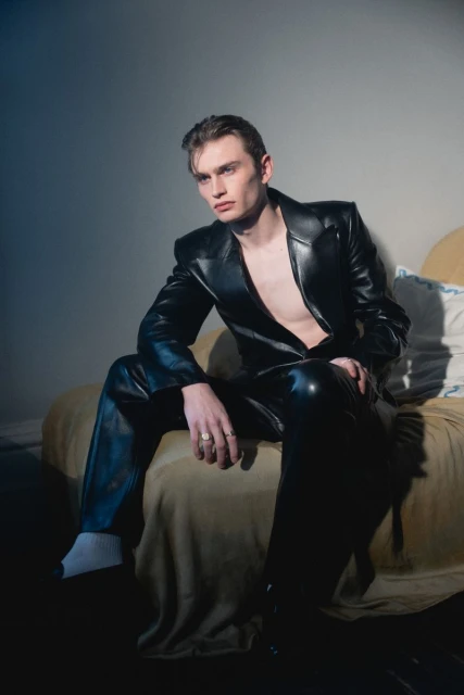 Fashionable young man in black leather suit poses on bed with yellow blanket and white pillows in studio lighting with grey background.