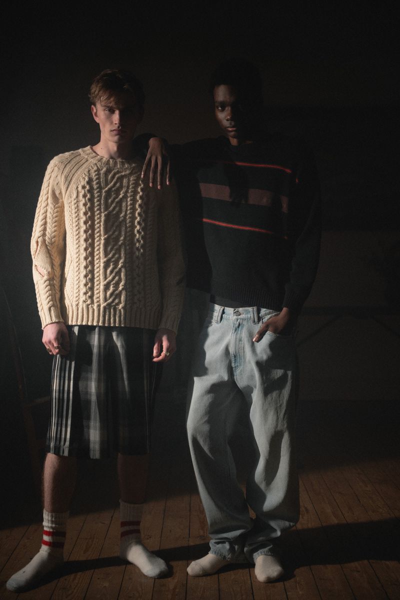 Two young adult men pose indoors under moody lighting, wearing casual sweaters, shorts, and jeans. A fashion-focused, visually striking image.