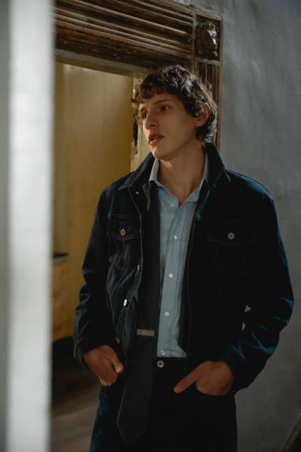 Portrait of a young adult male in a denim jacket and dress pants, looking to the side in an indoor setting with neutral lighting.
