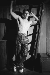 Shirtless male model posing against wooden walls with a ladder wearing dark trousers in a monochrome editorial photograph