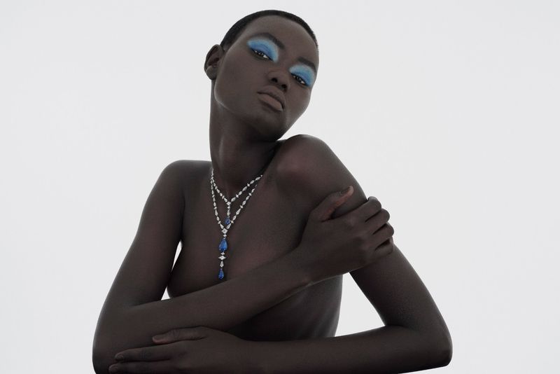 Dark-skinned woman with blue eyeshadow poses with necklace against a white backdrop in a beauty fashion studio portrait.