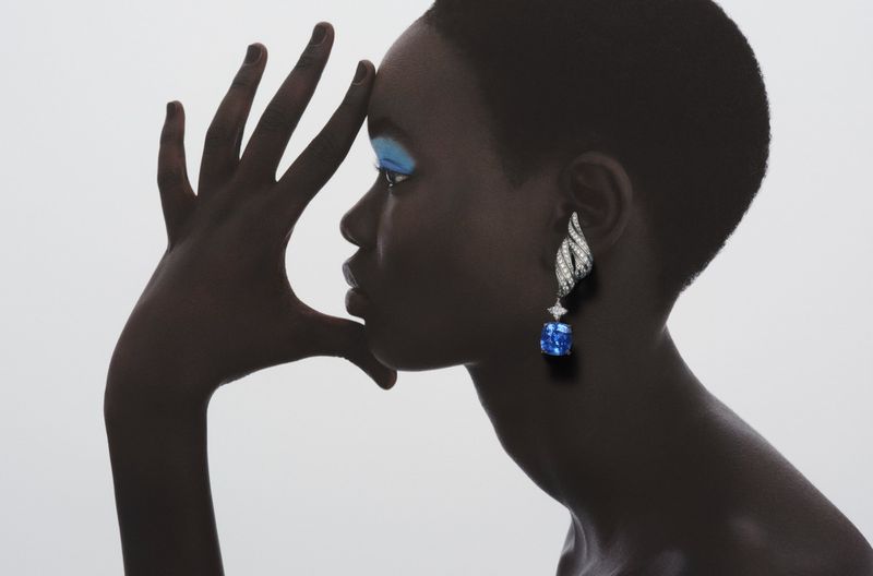 Close-up of a dark-skinned woman wearing a blue gemstone earring with a hand near her face against a white background.