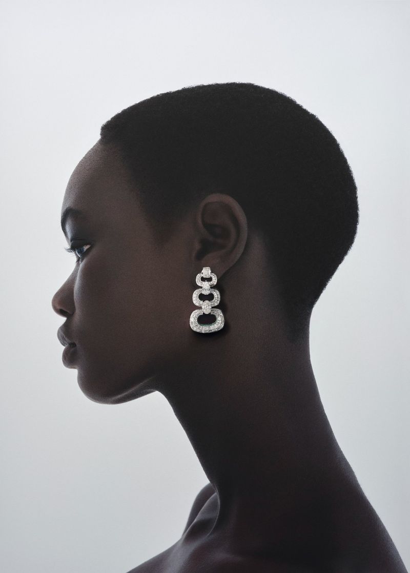 Profile view of a dark-skinned woman with a shaved head wearing dangling square earrings in a clean, white studio setting.