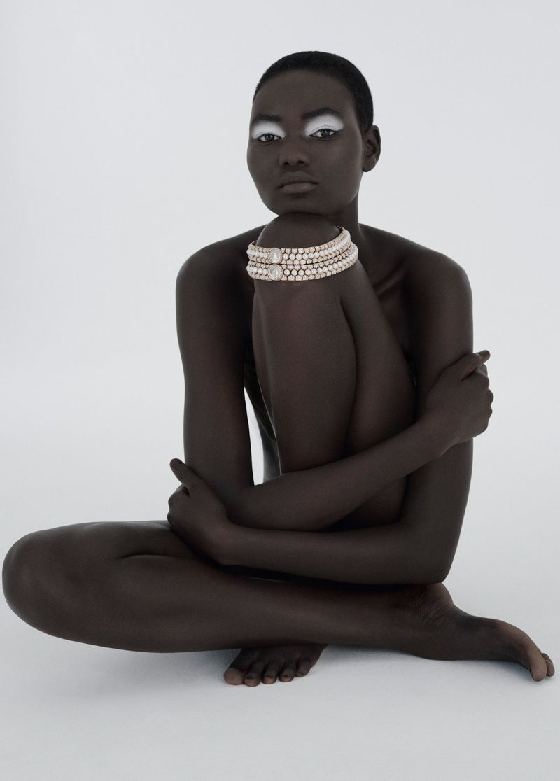 Elegant studio shot of a bald Black woman with white eyeshadow and pearl bracelets, seated against a bright white backdrop.