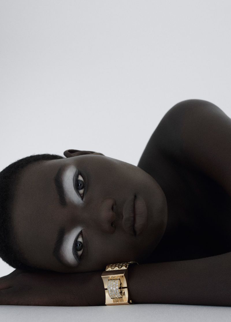 Close-up shot of a dark-skinned woman with white makeup around her eyes, laying down wearing a golden bracelet, against a white backdrop.