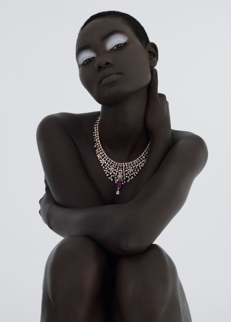 Striking portrait of a dark-skinned model with white makeup around the eyes, wearing an ornate diamond and ruby necklace against a bright white background.