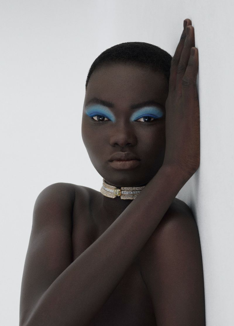 Striking studio shot of a bare-shouldered Black woman with blue eyeshadow and a choker, her hand against a white wall, gazing intensely.
