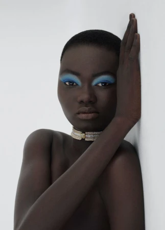Striking studio shot of a bare-shouldered Black woman with blue eyeshadow and a choker, her hand against a white wall, gazing intensely.