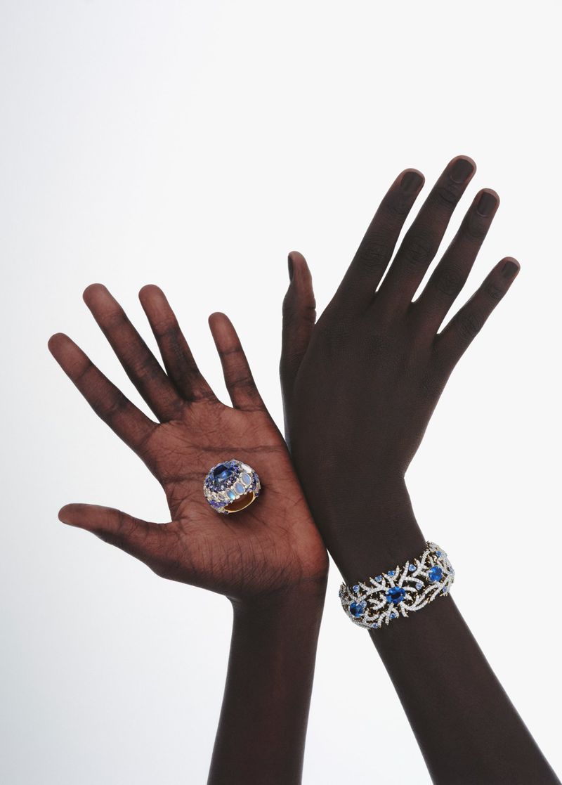 A dark-skinned person presents a jeweled ring on their palm alongside a jeweled bracelet, set against a bright white backdrop.