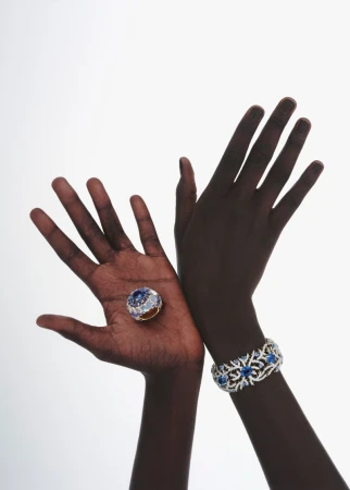 A dark-skinned person presents a jeweled ring on their palm alongside a jeweled bracelet, set against a bright white backdrop.