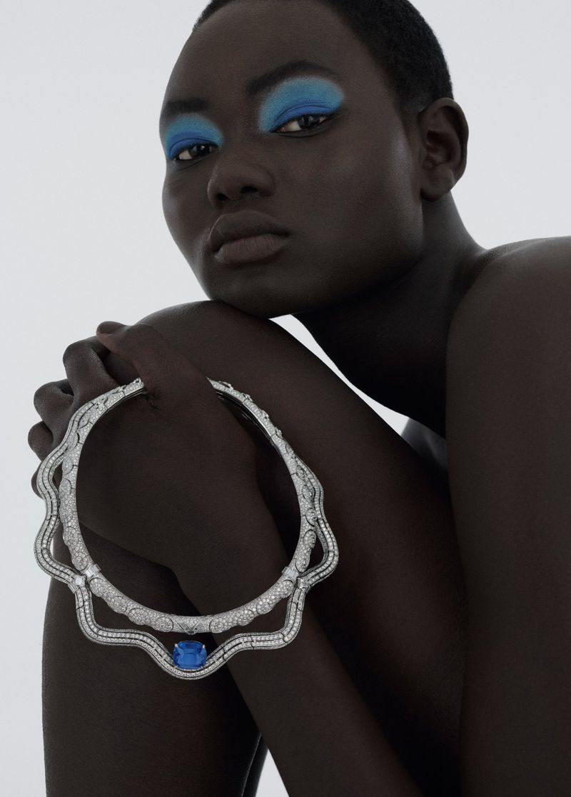Close-up shot of a dark-skinned female model with bright blue eyeshadow, holding an ornate silver necklace with blue gem in studio.