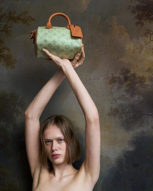 Model holding up a patterned green leather handbag with tan leather handles against a textured dark painterly background.