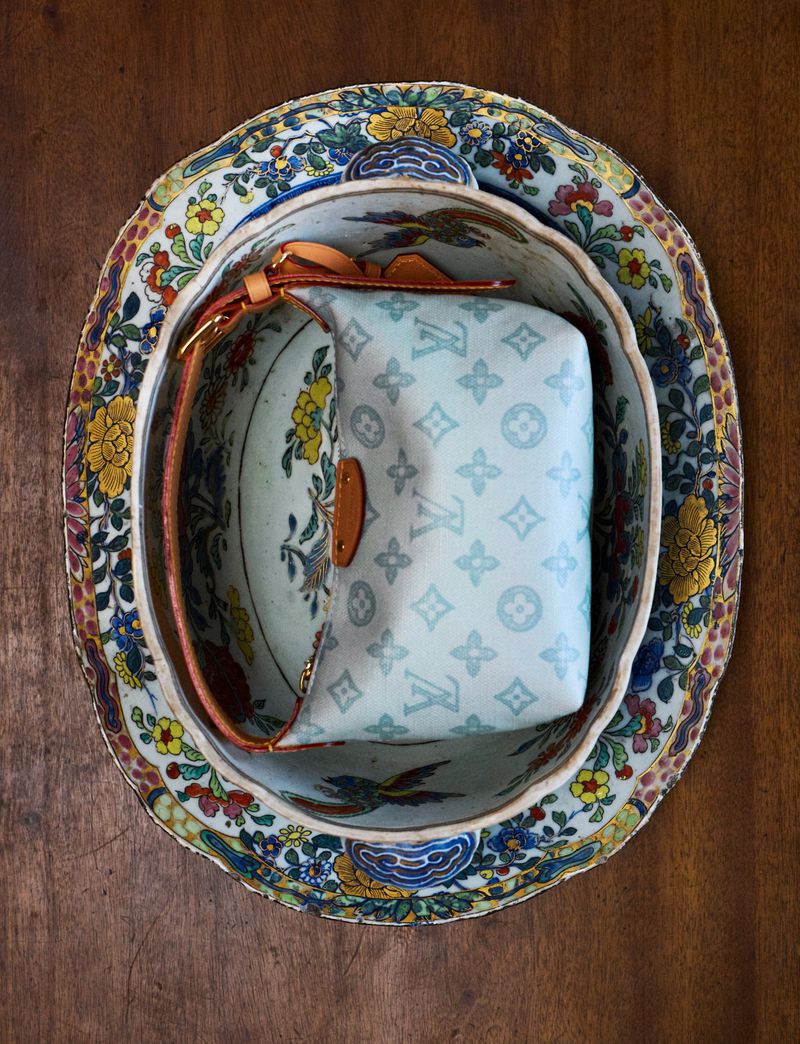 Overhead shot of a light blue Louis Vuitton handbag with brown leather accents inside of a decorative, vintage plate set on a wood surface.